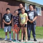 Native american children and adult stand beside new water spigot outside rural home