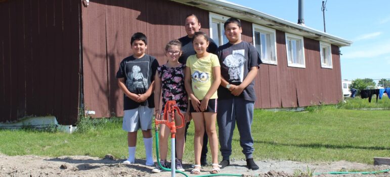 Native american children and adult stand beside new water spigot outside rural home