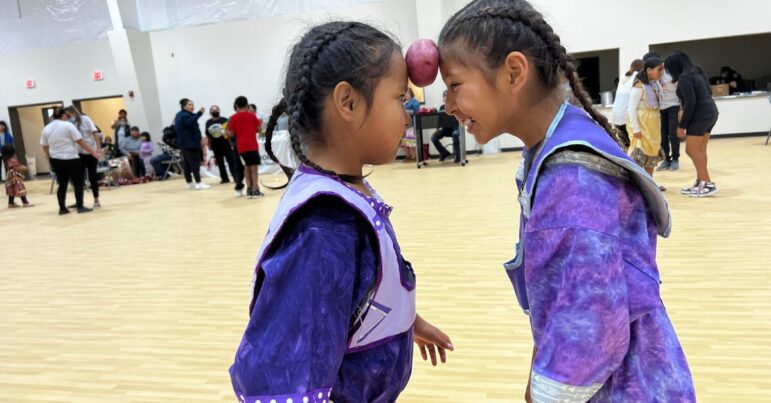 wo Native American girls in traditional regalia playing a balancing game at Oyate Ta Kola Ku Community Center.