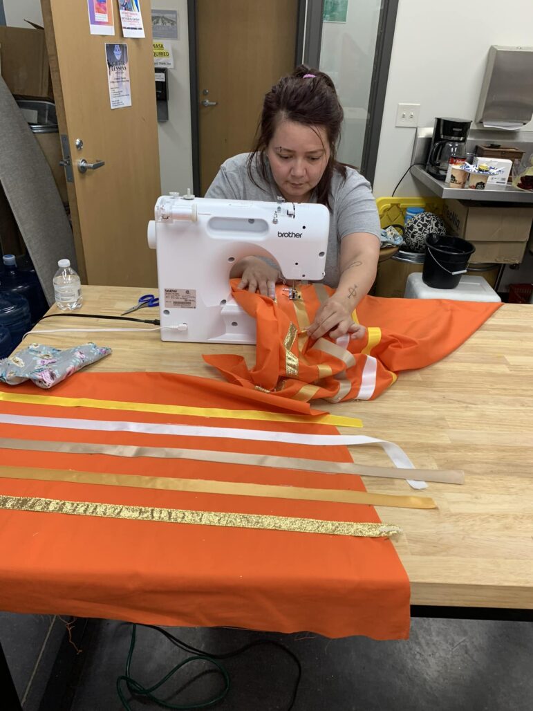 Woman sewing a Lakota ribbon skirt with orange fabric and decorative ribbons.