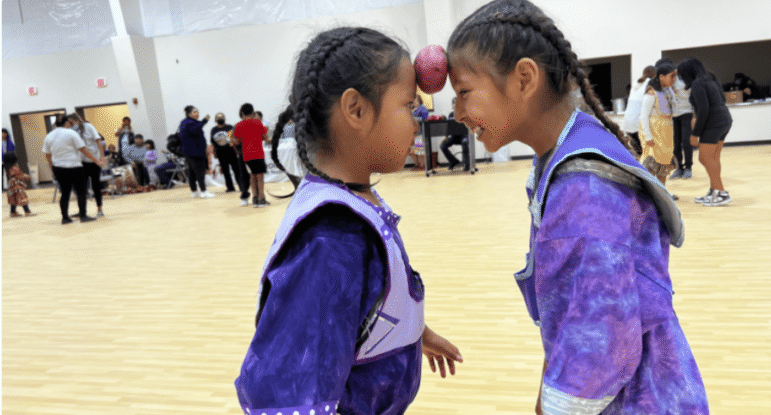 Two girls smiling as they balance a potato between their foreheads while doing the potato dance at a community center activity.