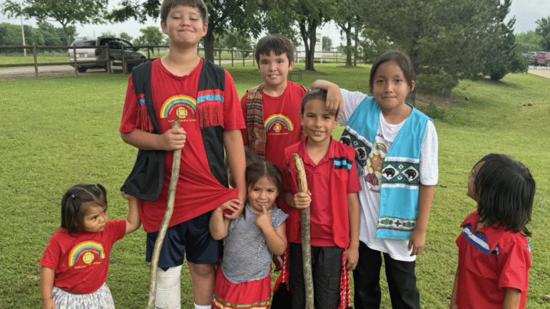 A group of smiling Yuchi children from the language immersion program stand together outdoors.