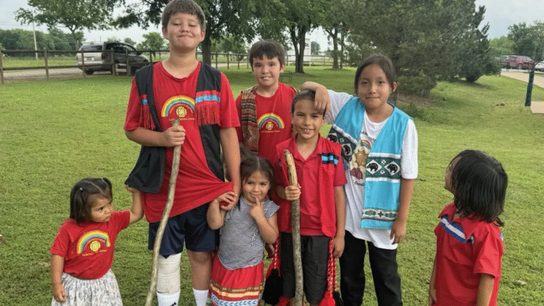 A group of seven Yuchi children of various ages smiling together on a grassy field in Sapulpa, Oklahoma.