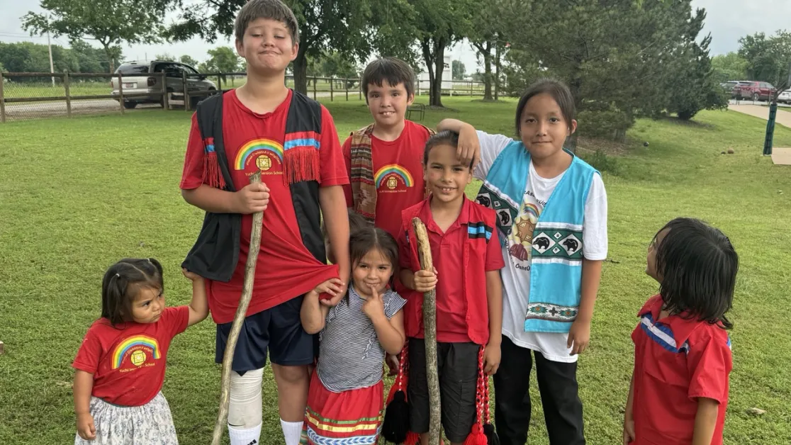 A group of seven Yuchi children of various ages smiling together on a grassy field in Sapulpa, Oklahoma.
