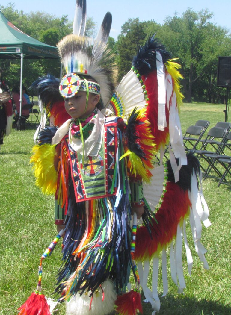 Young Native American boy in colorful powwow regalia with feathered bustles, standing outdoors at Running Strong event.