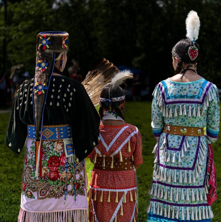 Honoring Indigenous legacies, three Native women in vibrant, traditional regalia walk at a cultural gathering.