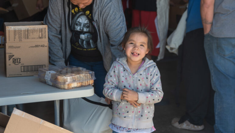 A smiling child stands by a table of food and snacks provided by the Running Strong food security program.