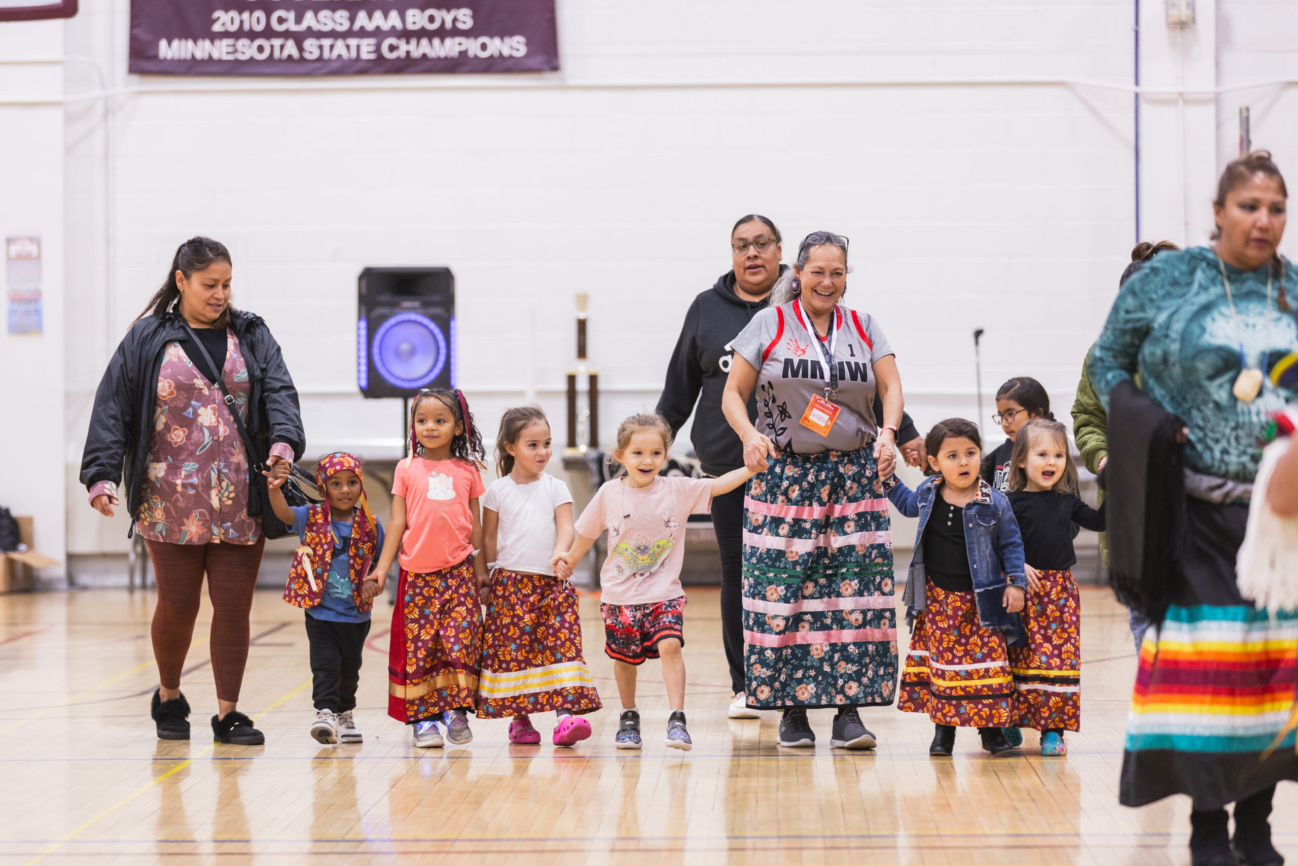 Adults and children in traditional ribbon skirts walk hand-in-hand in a gym.