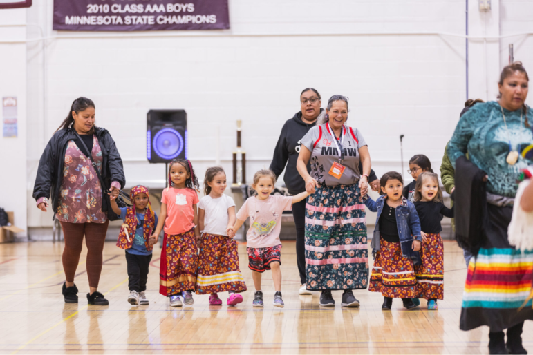 Indigenous women and young girls wearing traditional ribbon skirts holding hands and walking across a gymnasium floor.