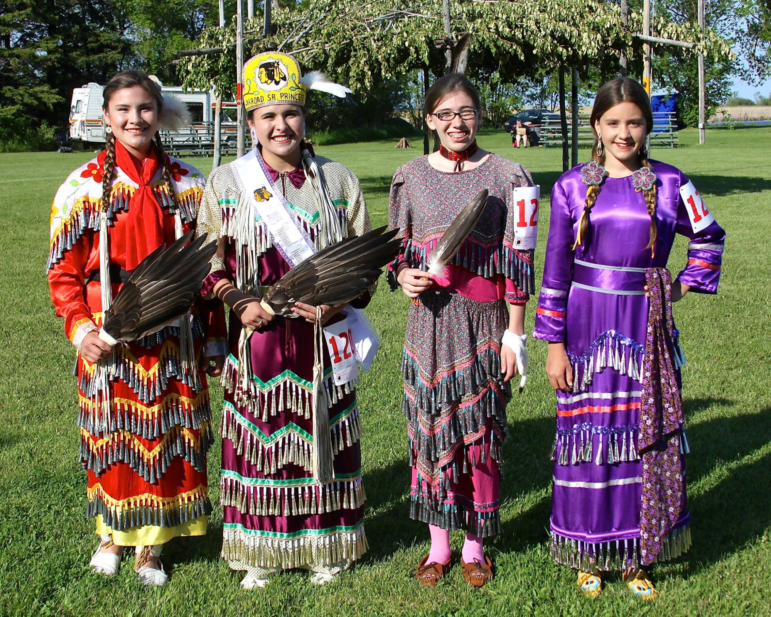 Four young Native American women wearing colorful traditional dresses and holding feather fans posing together outdoors.