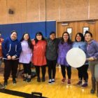 Community members in regalia standing together with a hand drum at an indoor gathering.