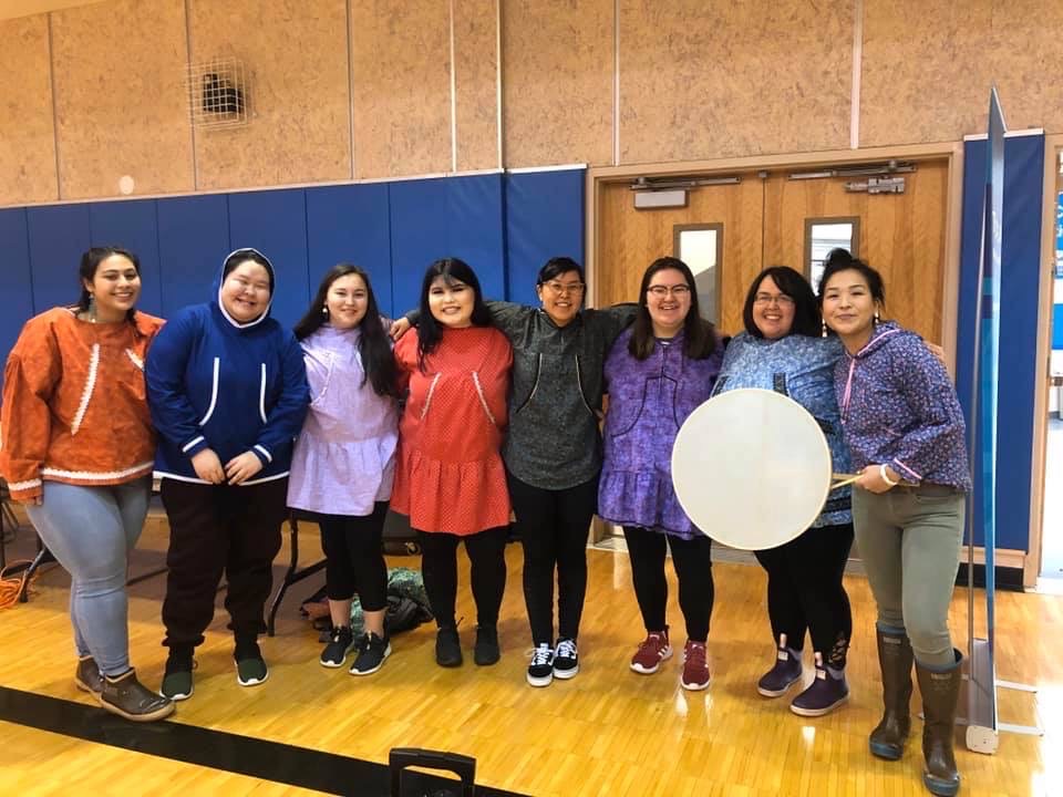 Community members in regalia standing together with a hand drum at an indoor gathering.