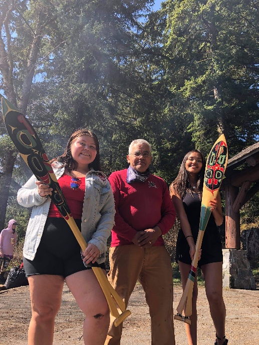 Three community members standing outdoors, with two holding decorated paddles.
