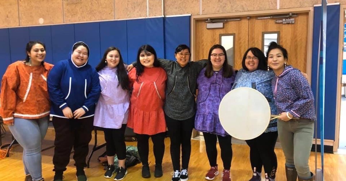 Community members in regalia standing together with a hand drum at an indoor gathering.