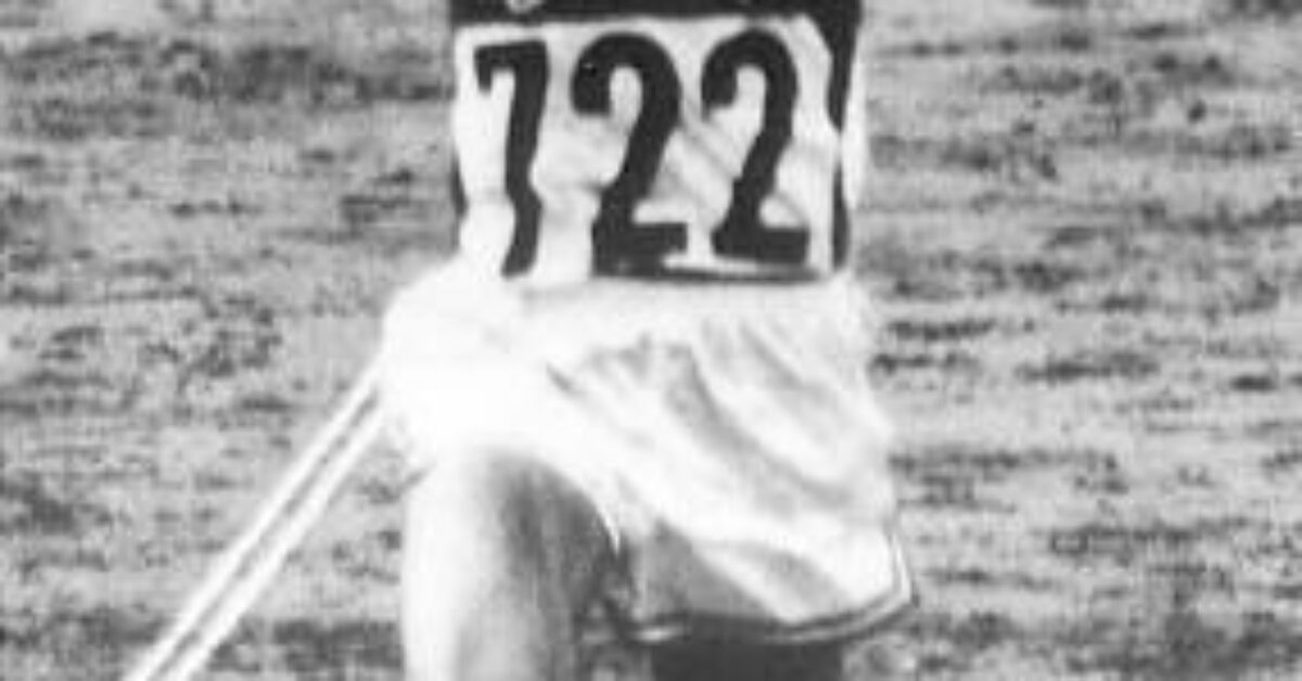 Billy Mills (USA 722) crosses the finish line with arms raised, winning the 10,000m gold medal at the 1964 Tokyo Olympics.
