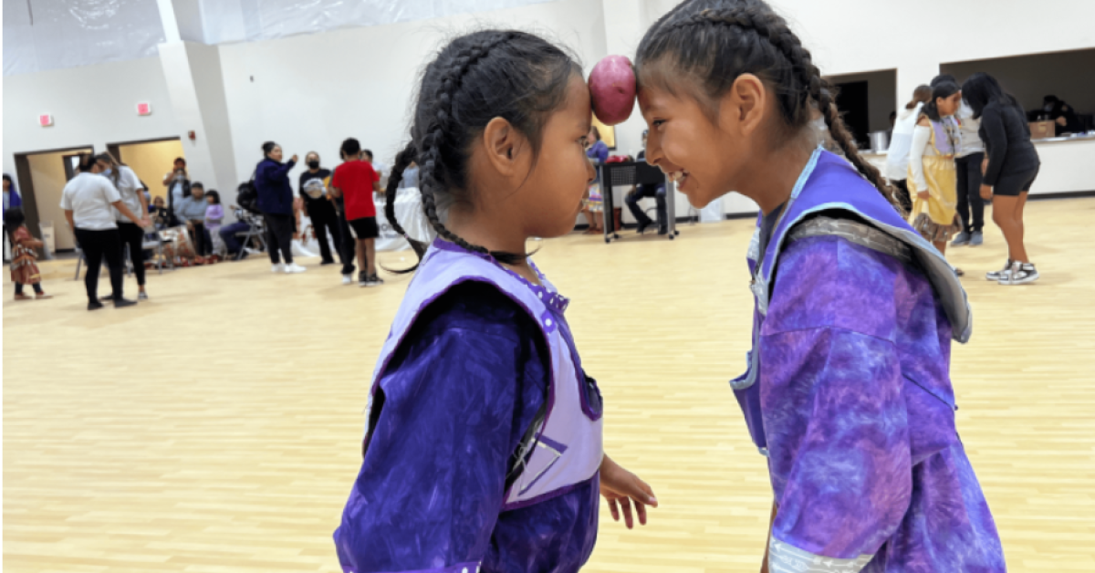 Two girls smiling as they balance a potato between their foreheads while doing the potato dance at a community center activity.