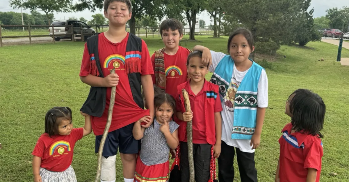 A group of seven Yuchi children of various ages smiling together on a grassy field in Sapulpa, Oklahoma.